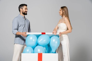 Neşeli ve şık hamile bir kadın kocasına bakarken cinsiyet ayrımında mavi balonlar olan büyük hediye kutusunu açıyor. Gri renkte izole edilmiş sürpriz bir parti.