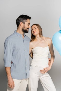 Gülümseyen ve şık sakallı bir adam hamile karısına sarılıp bakarken mavi balonların yanında duruyor. Gri arka planda sürpriz bir parti var.