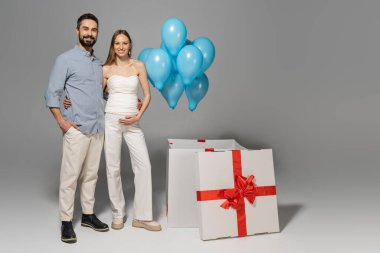 Tam uzunlukta neşeli ve modaya uygun ebeveynler kameraya sarılıp bakarken büyük hediye kutusunun yanında ve cinsiyet ayrımı sırasında mavi balonlar gri arka planda sürpriz bir parti olduğunu gösteriyor.