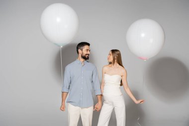 Positive and trendy expecting parents holding hands and festive balloons during celebration and gender reveal surprise party on grey background, expecting parents concept clipart