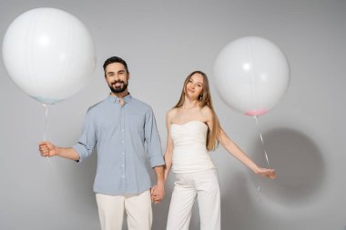 Gülümseyen ve stil sahibi aileler el ele tutuşurken ve kameraya bakarken beyaz balonlar beklerken gri arkaplanda sürpriz bir parti olduğunu gösteriyor.
