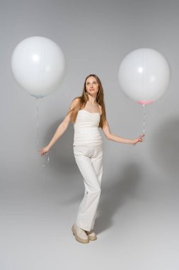 Full length of trendy and pregnant woman looking away while holding white festive balloons during celebration and gender reveal surprise party on grey background, fashionable pregnancy attire clipart