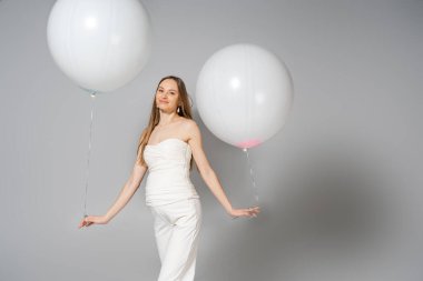 Positive and fashionable pregnant woman looking at camera while holding white festive balloons during gender reveal surprise party on grey background, fashionable pregnancy attire clipart