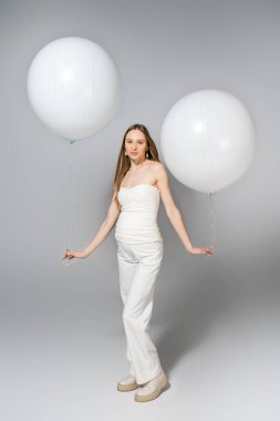 Full length of trendy and fair haired pregnant woman holding white balloons and looking at camera during gender reveal surprise party on grey background, fashionable pregnancy attire clipart