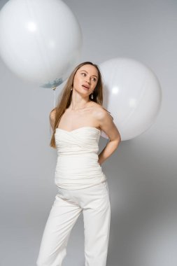 Düşünceli ve modaya uygun hamile bir kadın elinde şenlikli beyaz balonlar tutarken ve cinsiyet ayrımında ayakta dururken gri arka planda sürpriz bir parti veriyor.