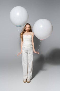 Full length of stylish and shocked expecting mother lookin at camera while holding white festive balloons during gender reveal surprise party on grey background, fashionable pregnancy attire clipart