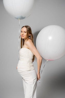 Gülümseyen ve modaya uygun hamile kadın kameraya bakıyor ve kutlama sırasında elinde beyaz balonlar tutuyor ve cinsiyet ayrımcılığı sırasında sürpriz parti gri arka planda, modaya uygun hamilelik kıyafetleri giyiyor.