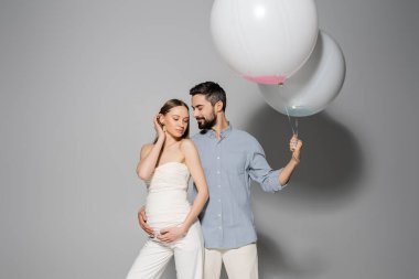 Gülümseyen ve sakallı bir adam şık hamile eşine sarılıyor, cinsiyet partisi sırasında balon tutuyor ve gri arka planda kutlama yapıyor, ebeveyn kavramı bekliyor, erkek ya da kız. 