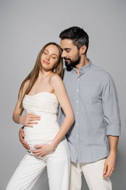 Sakallı ve şık bir adam gri arka planda birlikte dururken rahat ve modaya uygun hamile bir kadına sarılıp ebeveyn konsepti, bebek çarpması, karı koca... 