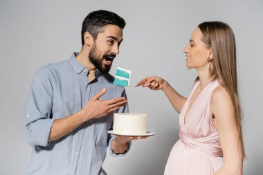 Smiling pregnant woman in pink dress holding blue cake near husband with open mouth during gender party celebration on grey background, expecting parents concept, it`s a boy  clipart