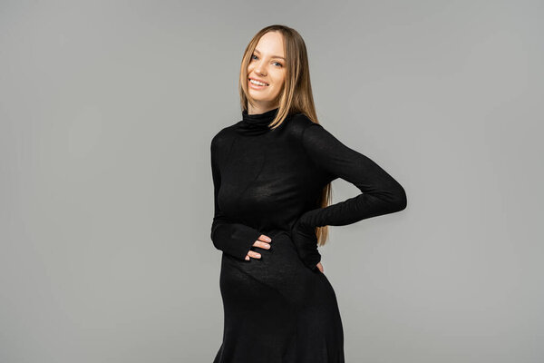 Smiling fair haired and pregnant woman with everyday makeup in stylish black dress touching belly and looking at camera while standing isolated on grey, growing new life concept