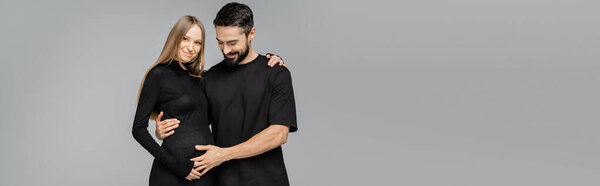 Cheerful bearded man in black t-shirt touching belly of fair haired and pregnant stylish wife in dress and hugging isolated on grey, growing new life concept, copy space, banner 