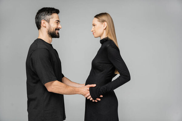 Side view of positive bearded man in t-shirt touching belly of pregnant wife in stylish black dress and looking at each other while standing isolated on grey, growing new life concept