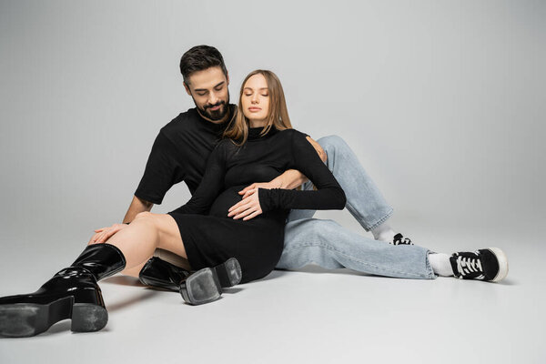 Bearded man in jeans and t-shirt touching belly of fair haired and stylish pregnant wife in black dress while sitting on grey background, new beginnings and parenting concept 