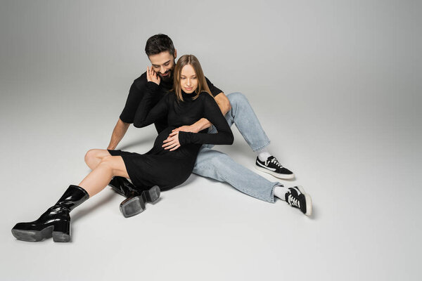 Smiling bearded man in jeans and t-shirt hugging fashionable pregnant woman in dress while sitting on grey background, new beginnings and parenting concept, husband and wife 