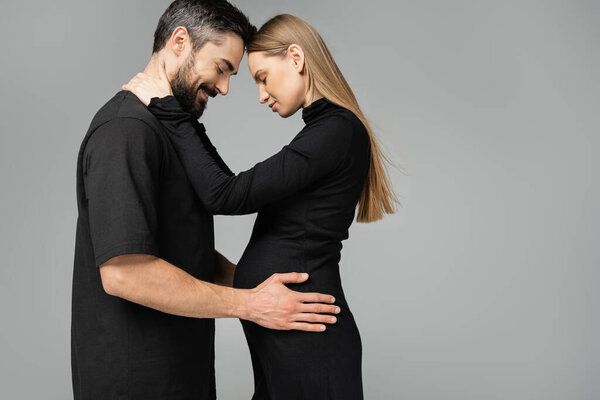 Side view of stylish and fair haired pregnant woman in black dress hugging cheerful husband with closed eyes and standing isolated on grey, new beginnings and anticipation concept  