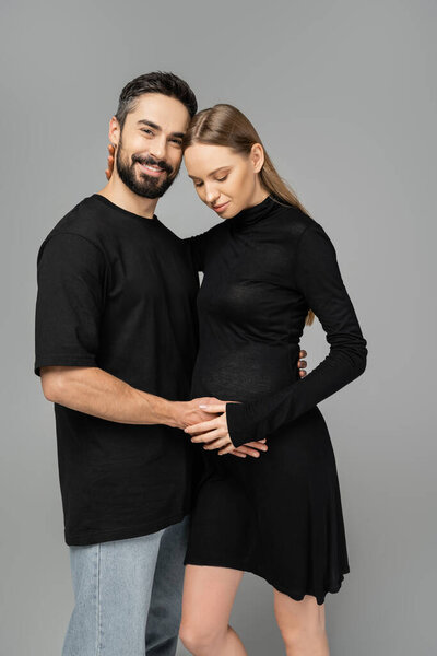 Positive and bearded man in jeans and t-shirt hugging trendy pregnant wife in black dress and looking at camera isolated on grey, new beginnings and anticipation concept  