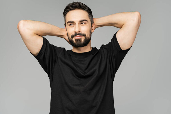 Joyful bearded and brunette man in black t-shirt touching head and looking away while standing isolated on grey, masculine beauty concept, confident and charismatic 