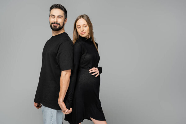Positive bearded man holding hand of stylish pregnant wife in black dress and standing back to back isolated on grey, new beginnings and parenting concept  