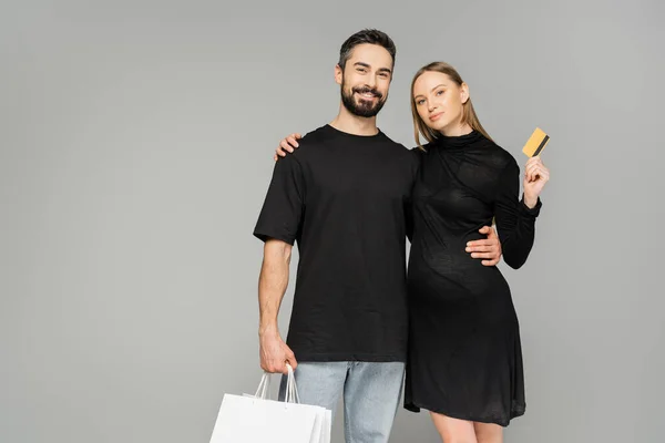 满脸胡须的男人拿着购物袋 一边拿着信用卡拥抱怀孕的妻子 一边看着相机 一边孤身一人站在灰色 新开端和养育孩子的观念上 — 图库照片