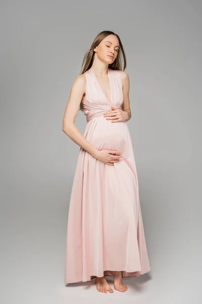 stock image Full length of barefoot and fair haired pregnant woman in elegant pink dress touching belly and standing with closed eyes on grey background, elegant and stylish pregnancy attire