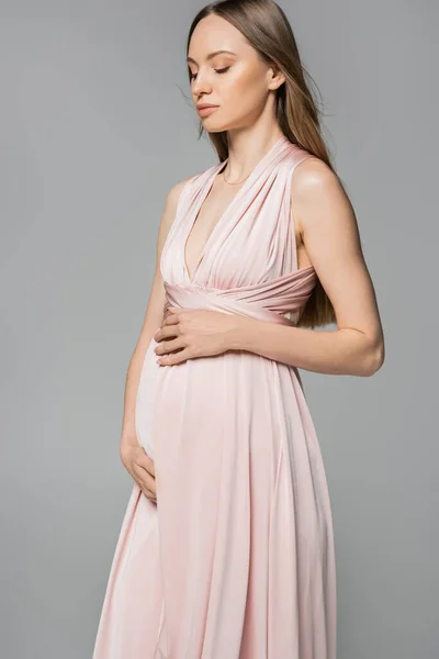Fair haired and pregnant woman in pink dress touching belly and looking down while standing isolated on grey, elegant and stylish pregnancy attire, sensuality, mother-to-be