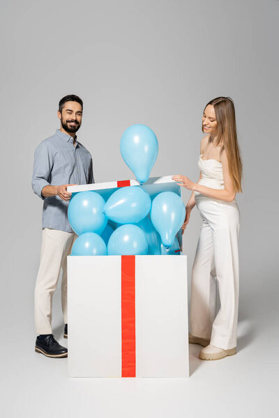 Cheerful and stylish pregnant woman opening big gift box with blue balloons near husband during celebration and gender reveal surprise party on grey background, it`s a boy 