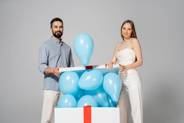 Positive and stylish expecting parents looking at camera while opening big gift box with blue balloons during gender reveal surprise party and celebration isolated on grey, it`s a boy 