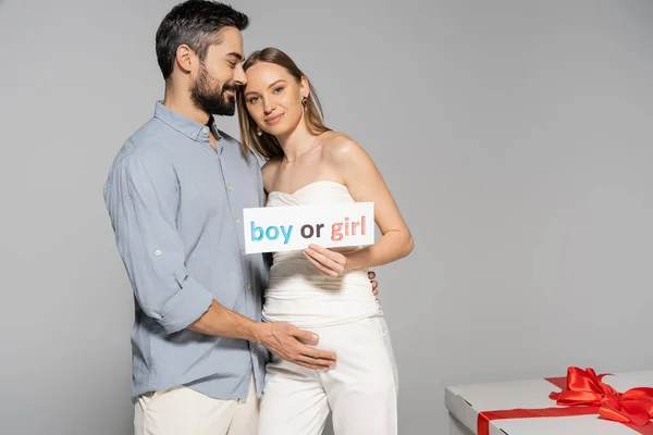 Smiling pregnant woman holding card with boy or girl lettering near stylish husband and big gift box during gender reveal surprise party isolated on grey, expecting parents concept