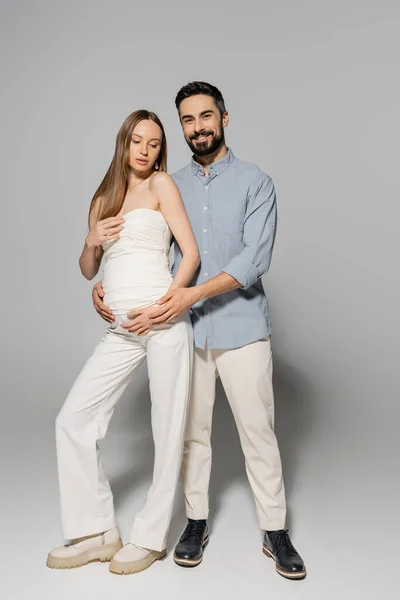 前向きな髭を生やした男がスタイリッシュで妊娠中の女性を抱きかかえてカメラを見ながらグレーの背景に立ち 両親のコンセプト 赤ちゃんのバンプ 夫と妻を期待 — ストック写真