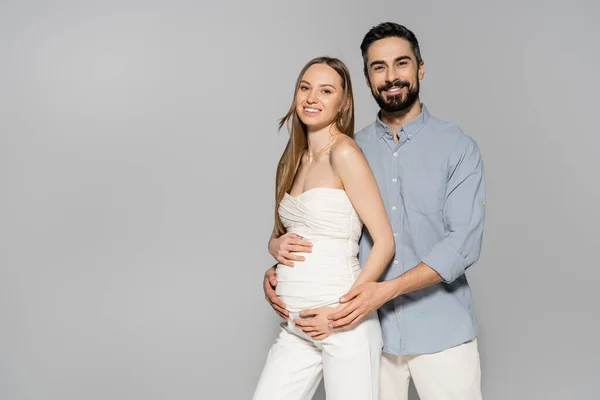 笑顔と髭を生やした男はファッショナブルな妊娠中の女性の腹を抱き カメラを見ながら一緒にグレーに隔離され 両親の概念 赤ちゃんのバンプ 夫と妻を期待 — ストック写真