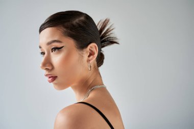Genç ve büyüleyici Asyalı kadın portresi, esmer saçlı, kalın makyajlı, şık saç stili gümüş aksesuarlarla poz veren ve gri arka planda, şık bahar, moda fotoğrafçılığı.