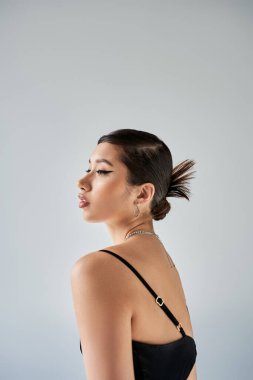 İlkbahar stili koyu makyajlı Asyalı kadın, esmer saçlı, modaya uygun saç stili, gümüş kolyeli, gri arka planda siyah ve zarif kayışlı elbise, bahar modası fotoğrafçılığı.