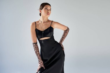 Bahar stili, esmer ve kalın makyajlı ifade edici Asyalı kadın. Siyah, zarif bir elbise giymiş, gri arka planda hayvan desenli eldivenler ve gümüş kolyeler takıyor.