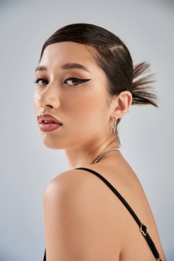 Genç ve çekici Asyalı kadın portresi anlamlı bakışlar, kalın makyaj, esmer saçlar, moda saç stili ve gri arka planda kameraya bakan gümüş aksesuarlar, bahar moda fotoğrafçılığı.