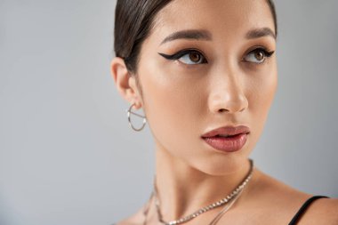Gümüş kolyeli ve küpeli Asyalı bir kadının portresi, esmer saçlı, cesur makyajlı ve etkileyici bakışları gri arka planda, moda fotoğrafçılığı, moda fotoğrafçılığı.