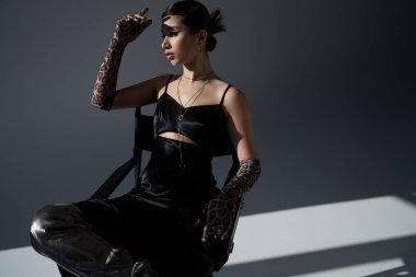 Genç Syle, bahar modası, siyah zarif elbiseli genç Asyalı kadın ve sandalyede oturan hayvan desenli eldivenler koyu gri arka plan, z nesli, gümüş kolyeler. 