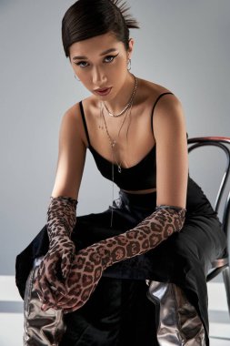 bahar moda fotoğrafçılığı, anlamlı bakışlı Asyalı kadın, hayvan desenli eldivenler, siyah kayış elbise ve gümüş kolyeler sandalyede oturuyor ve gri arka planda ışıklandırmalı kameraya bakıyor.