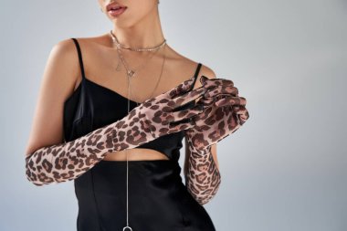 bahar moda fotoğrafçılığı, siyah kayışlı elbise ve gümüş kolyeli genç ve şık bir kadının gri arka planda hayvan desenli eldivenlerini çıkarması, z nesli.