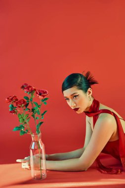 young and elegant asian woman in stylish neckerchief and dress, with bold makeup and brunette hair laying near glass vase with roses on red background with lighting, spring fashion, generation z clipart