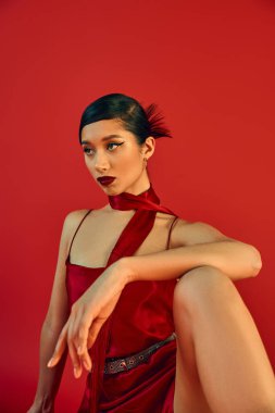 Cesur makyajlı genç ve kendinden emin Asyalı bir kadın, esmer saçlı ve etkileyici bakışları başka yöne bakan ve kırmızı arka planda zarif kayış elbisesiyle poz veren, bahar moda fotoğrafçısı.