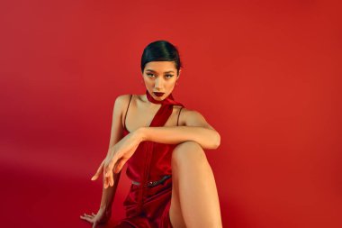 stylish spring concept, gen z fashion, young asian woman with brunette hair and bold makeup sitting in strap dress and scarf while looking at camera on red background clipart