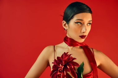 Çekici makyajlı, esmer ve etkileyici bakışlı Asyalı bir kadın. Kayışlı elbisesi ve atkısıyla bordo insanları tutuyor. Kırmızı arka planda, bahar moda fotoğrafçılığına bakıyor.