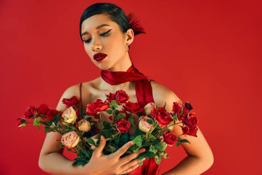 Çekici ve şık kolyeli Asyalı genç bir manken, esmer saçlı ve kalın makyajlı kırmızı arka planda taze güller, bahar moda fotoğrafçılığı.