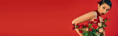 expressive and fashionable asian woman with bold makeup and brunette hair holding bouquet of roses and looking at camera on red background with copy space, youthful style, trendy spring, banner clipart