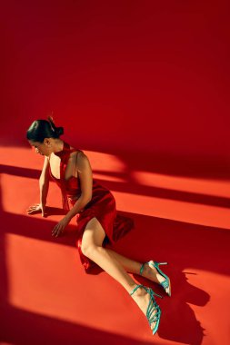 spring fashion photography, full length of graceful asian fashion model in strap dress, neckerchief and turquoise sandals sitting in lighting on red background, high angle view clipart