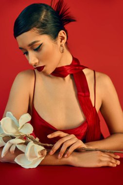 Moda elbisesi ve atkısı olan zarif ve genç Asyalı bir kadın, esmer saçlı ve kalın makyajlı, masada oturuyor ve kırmızı arka planda beyaz orkideye bakıyor, genel z moda, moda bahar konsepti