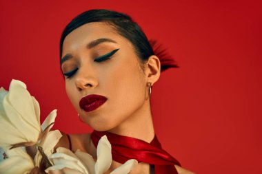 Esmer saçlı, seksi Asyalı mankenin portresi, koyu makyaj ve moda saç stili. Kırmızı arka planda beyaz çiçek açan orkidenin yanında gözler kapalı poz vermek, bahar modası, z nesli.