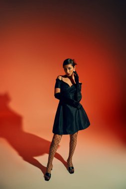full length of young asian woman with brunette hair, in long gloves, black cocktail dress and fishnet tights standing and looking away on red background with lighting effects, spring fashion concept clipart