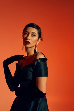 Siyah kokteyl elbisesi içinde şık ve çekici Asyalı manken, uzun eldivenler ve son moda küpeler omuza dokunuyor ve kırmızı ve turuncu arka plana bakıyor, bahar moda fotoğrafçılığı.